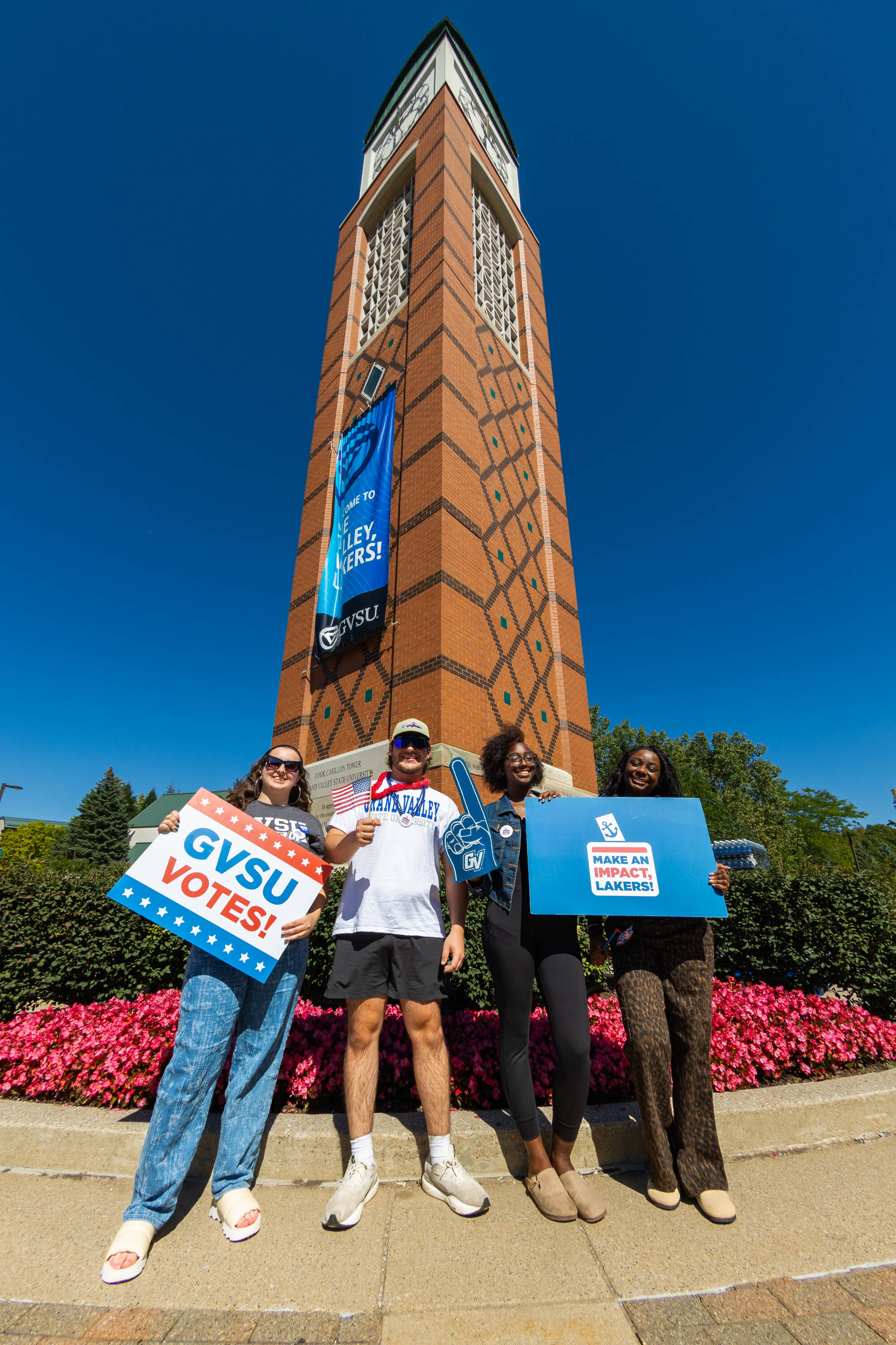 Four students standing by the clock tower and holding signs that say "GVSU Votes!" and "Make an impact, Lakers!"